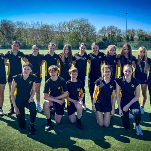 A group of students wearing navy and yellow sports kit pose together on an outdoor artificial pitch, arranged in two rows with some standing and others kneeling, with trees, fencing and a clear blue sky behind them
