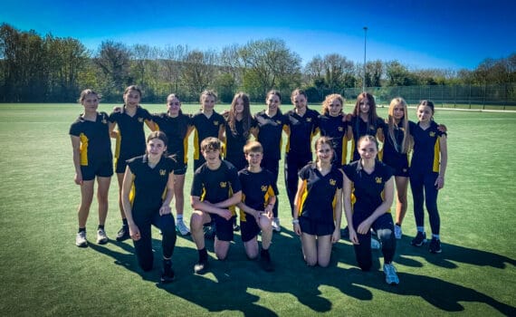 A group of students wearing navy and yellow sports kit pose together on an outdoor artificial pitch, arranged in two rows with some standing and others kneeling, with trees, fencing and a clear blue sky behind them