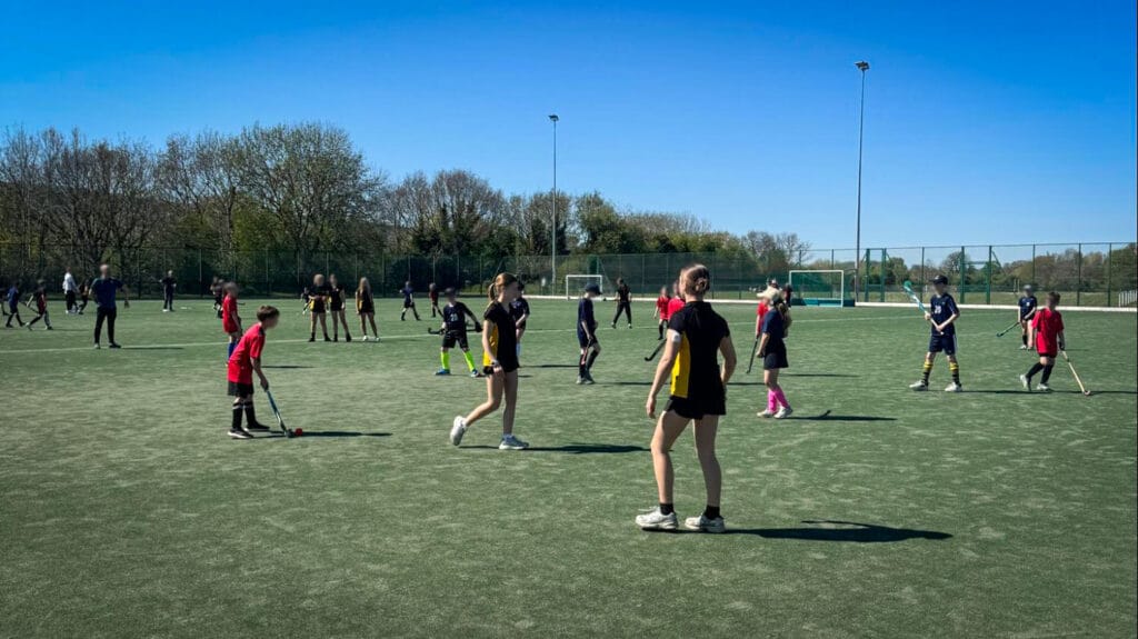 Students spread out across an outdoor pitch playing hockey, holding sticks and focusing on the ball as they prepare to pass or receive during a practice session