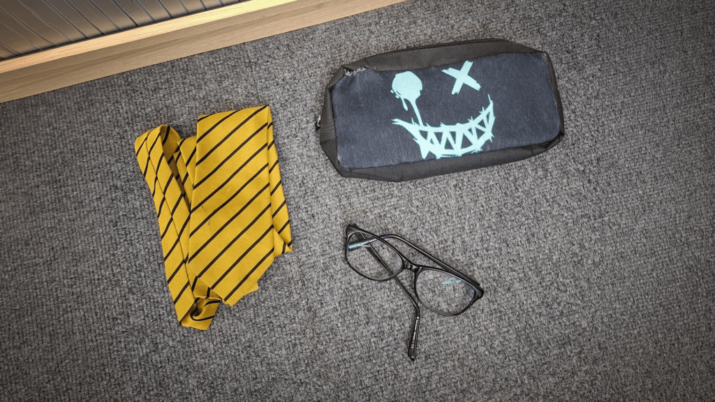 A yellow striped school tie, a pair of black-framed glasses and a grey pencil case with a stylised face design placed on a carpeted floor as part of the school’s lost property items