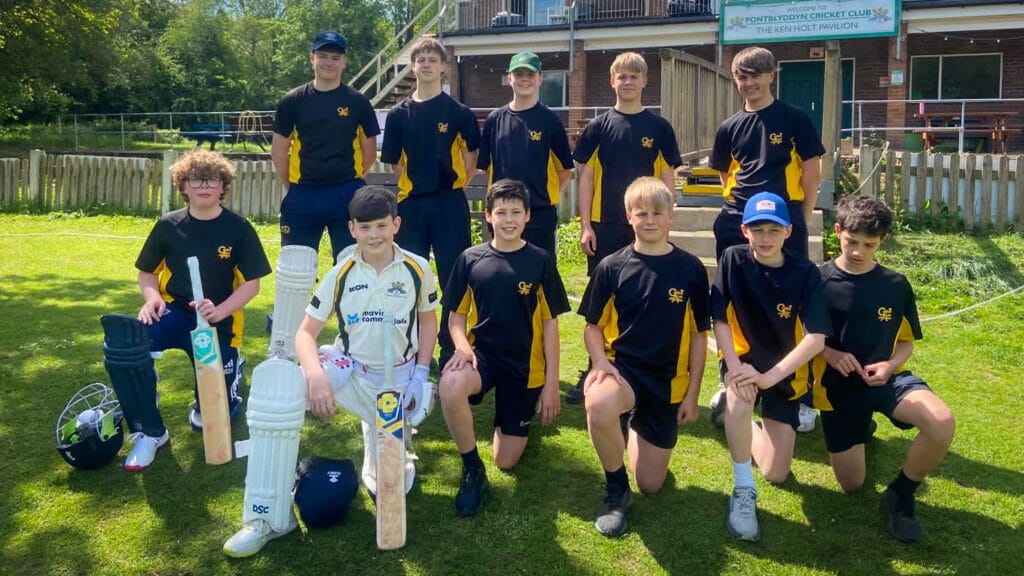 A group of students wearing navy and yellow sports kit pose together on a grassy cricket field outside a pavilion, with cricket bats, pads and helmets placed in front of them