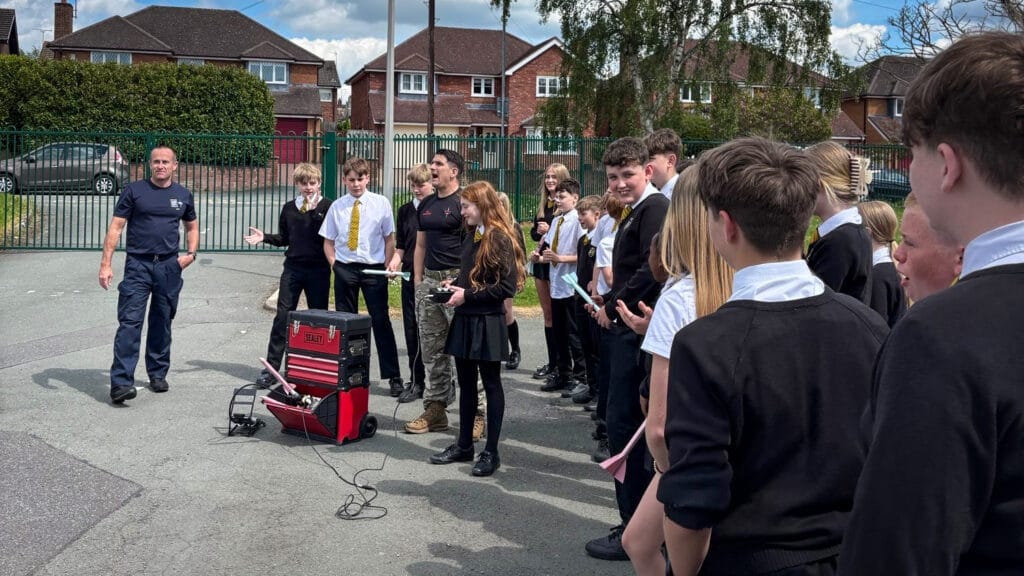 Students gathered in a school playground listen to a speaker speaking during an outdoor demonstration, with equipment cases and cables set up nearby