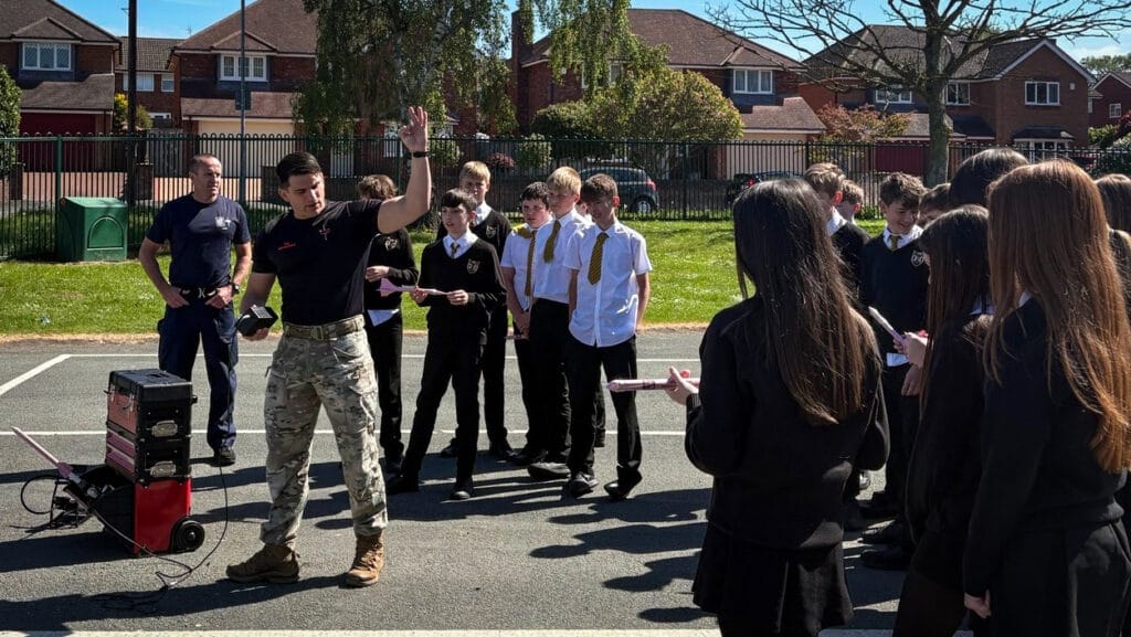 An instructor gestures while speaking to a group of students standing in a semi-circle on the playground during an outdoor safety activity