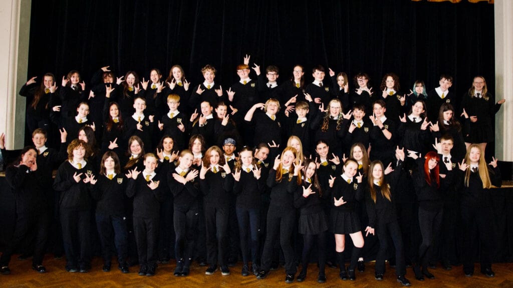 A large group of students dressed in school uniform pose together on a stage, smiling and making hand gestures in front of a dark theatre curtain
