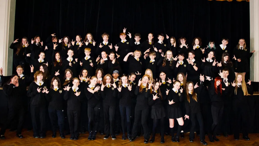 A large group of students dressed in school uniform pose together on a stage, smiling and making hand gestures in front of a dark theatre curtain
