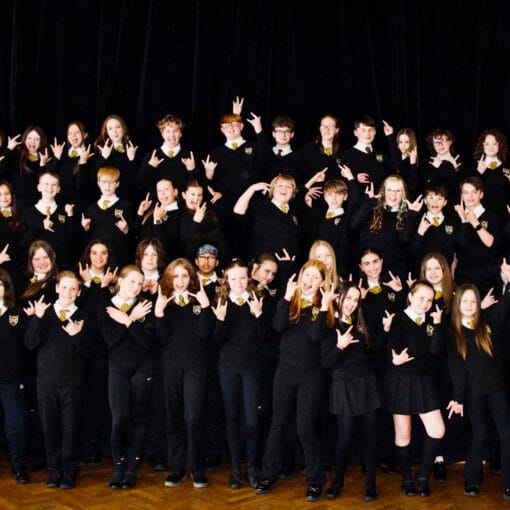 A large group of students dressed in school uniform pose together on a stage, smiling and making hand gestures in front of a dark theatre curtain