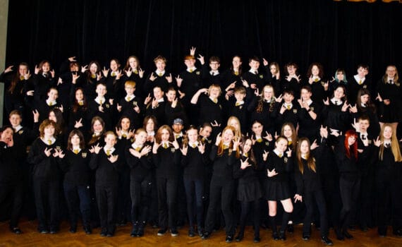 A large group of students dressed in school uniform pose together on a stage, smiling and making hand gestures in front of a dark theatre curtain
