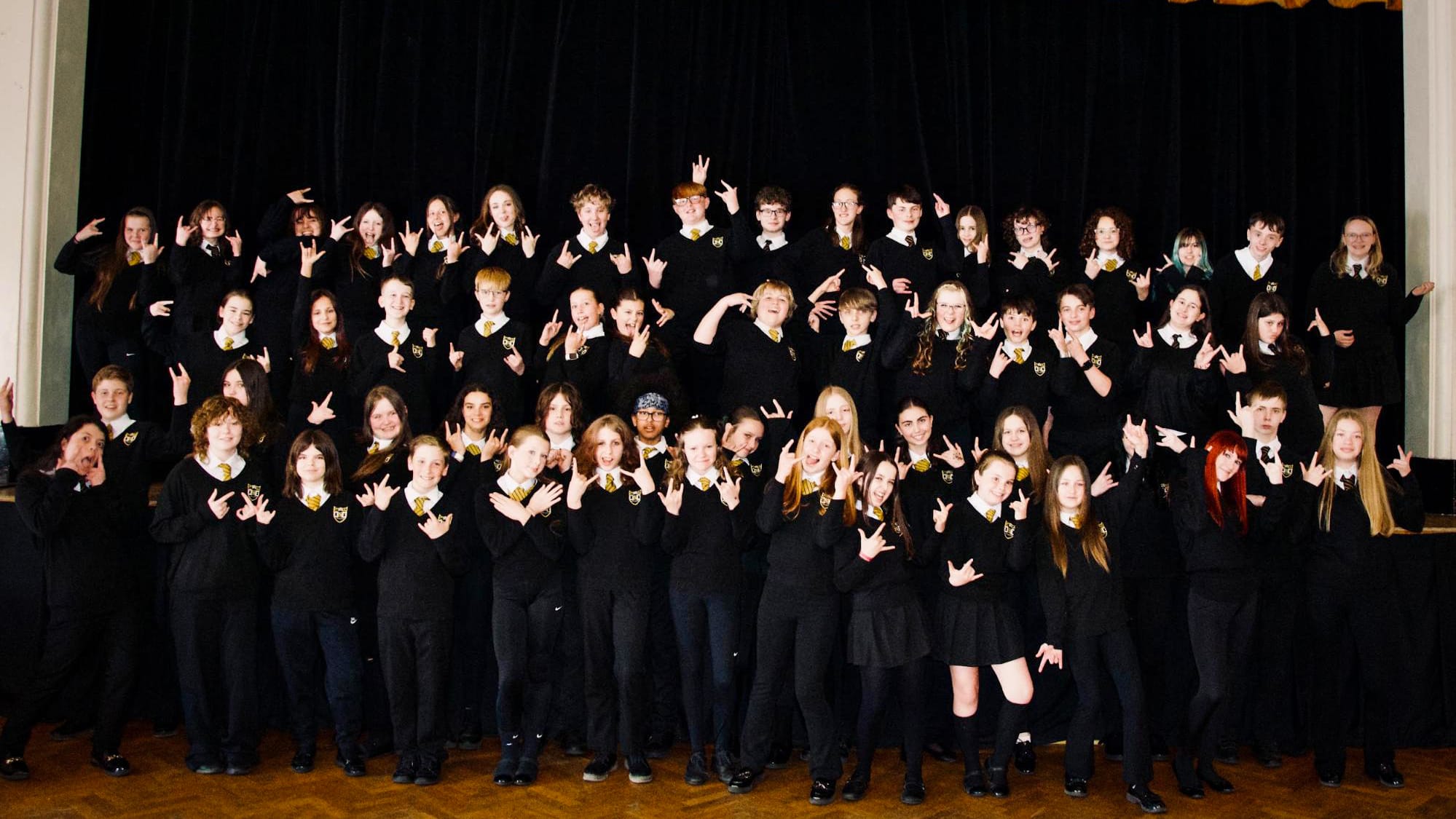 A large group of students dressed in school uniform pose together on a stage, smiling and making hand gestures in front of a dark theatre curtain