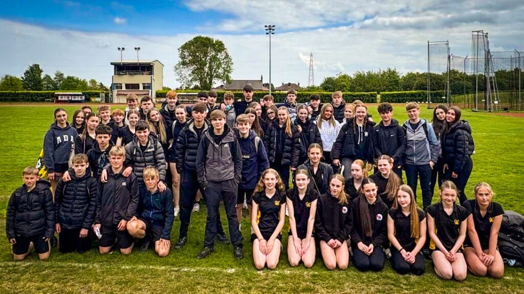 A large mixed group of students gathered together on a grassy athletics or sports field, with some standing and others kneeling during an outdoor school event