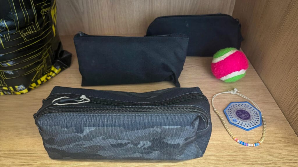 Several lost property items including black pencil cases, a pink and green tennis ball, a beaded bracelet and part of a black and yellow drawstring bag displayed on a wooden shelf
