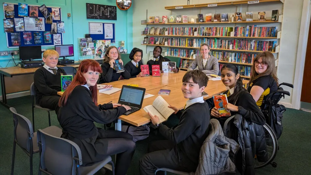 A group of students and a member of staff sit around tables in the school library, holding and reading books with shelves of colourful novels and displays in the background