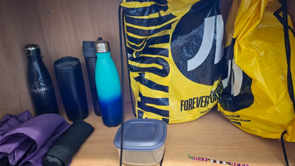 Several water bottles, yellow and black drawstring bags, an umbrella, a food container and a colourful beaded bracelet displayed together on a wooden shelf as part of the school’s lost property collection