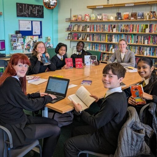A group of students and a member of staff sit around tables in the school library, holding and reading books with shelves of colourful novels and displays in the background