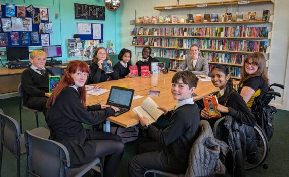 A group of students and a member of staff sit around tables in the school library, holding and reading books with shelves of colourful novels and displays in the background