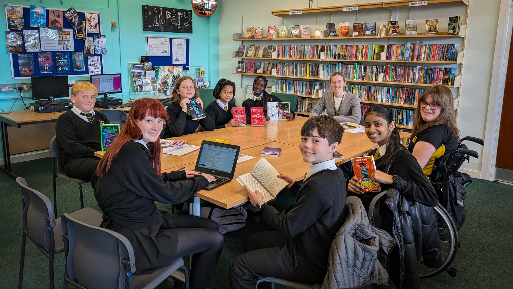 A group of students and a member of staff sit around tables in the school library, holding and reading books with shelves of colourful novels and displays in the background