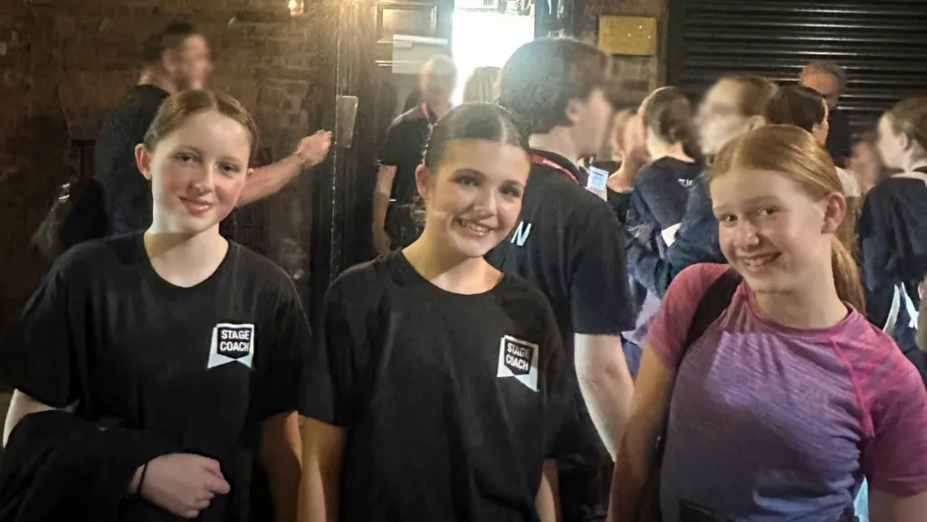 Three students smiling and posing together indoors while wearing Stage Coach theatre school clothing during a performance or rehearsal event