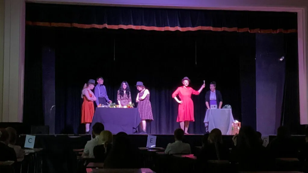 Students performing a scene on stage in costume in front of an audience seated at tables in a darkened hall