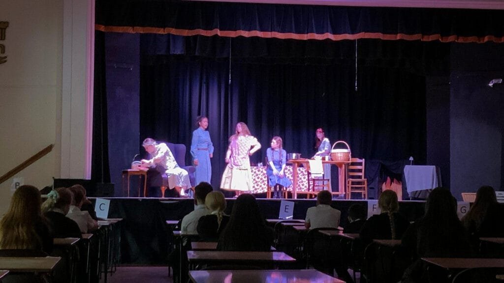 Students acting in a stage production with props and furniture on stage while an audience watches from tables in the foreground