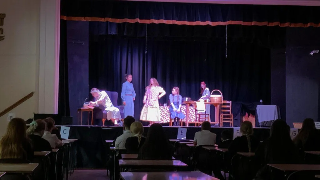 Students acting in a stage production with props and furniture on stage while an audience watches from tables in the foreground