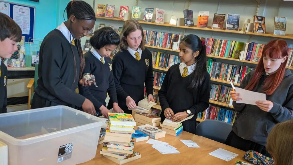 Students in school uniform sorting and organising books around tables in the school library with shelves of books behind them