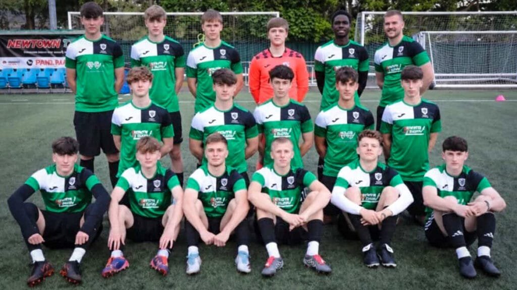 A football team in green, black and white kit pose together on an outdoor football pitch with coaches and goalposts in the background