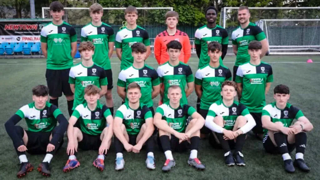A football team in green, black and white kit pose together on an outdoor football pitch with coaches and goalposts in the background