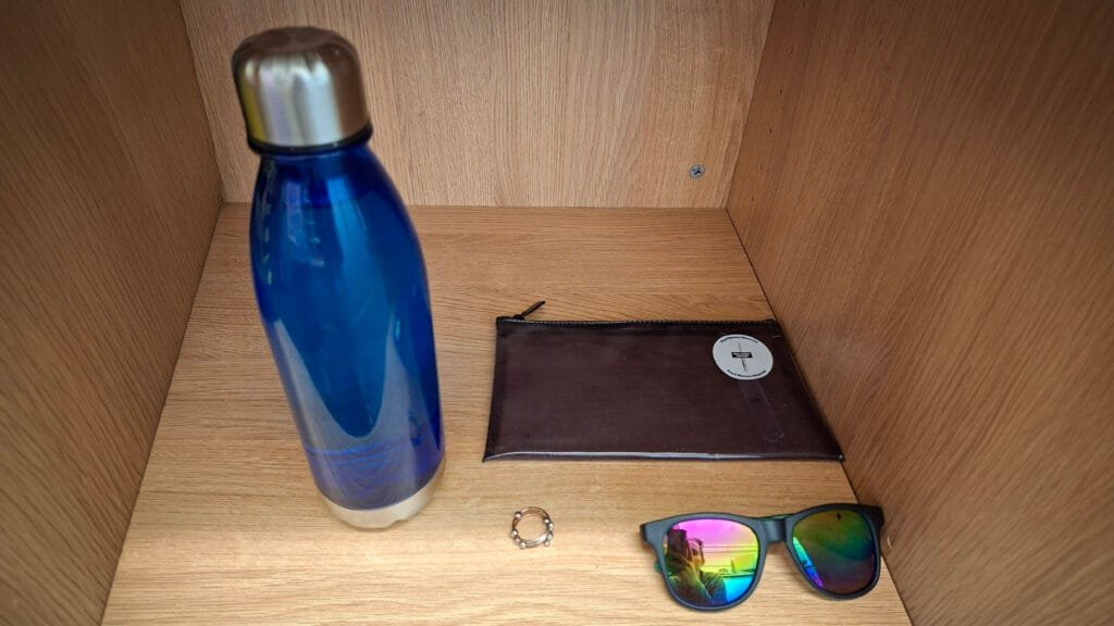 A blue metal water bottle, a brown zip pouch, a small ring, and a pair of colourful reflective sunglasses arranged on a wooden shelf.