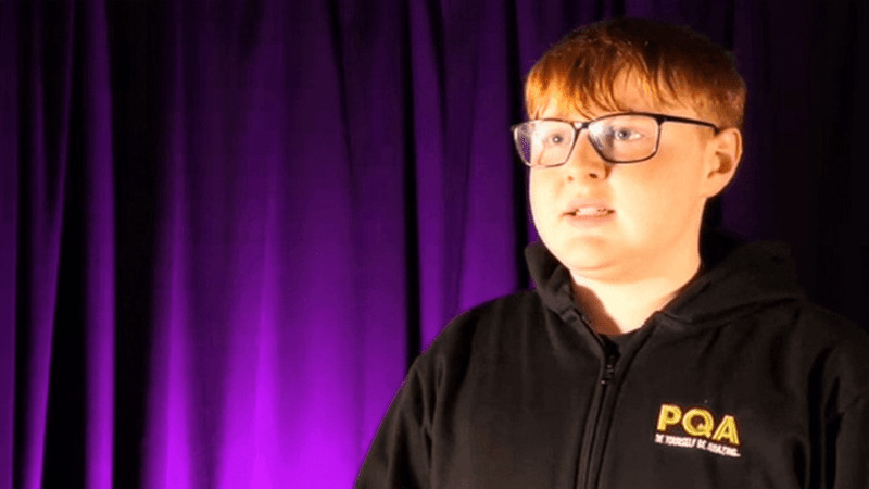 A student wearing glasses and a black hoodie with ‘PQA’ on the front, standing in front of a purple curtain