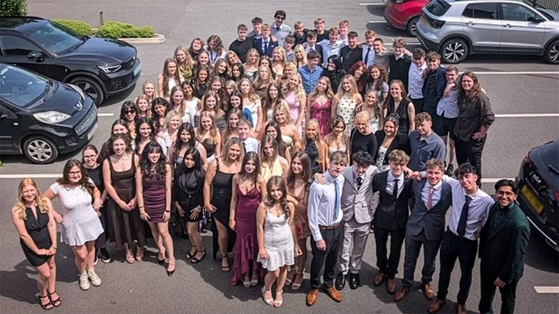 A large group of students dressed in formal attire, including dresses and suits, standing together in a car park with several parked cars around them, posed for a group photo
