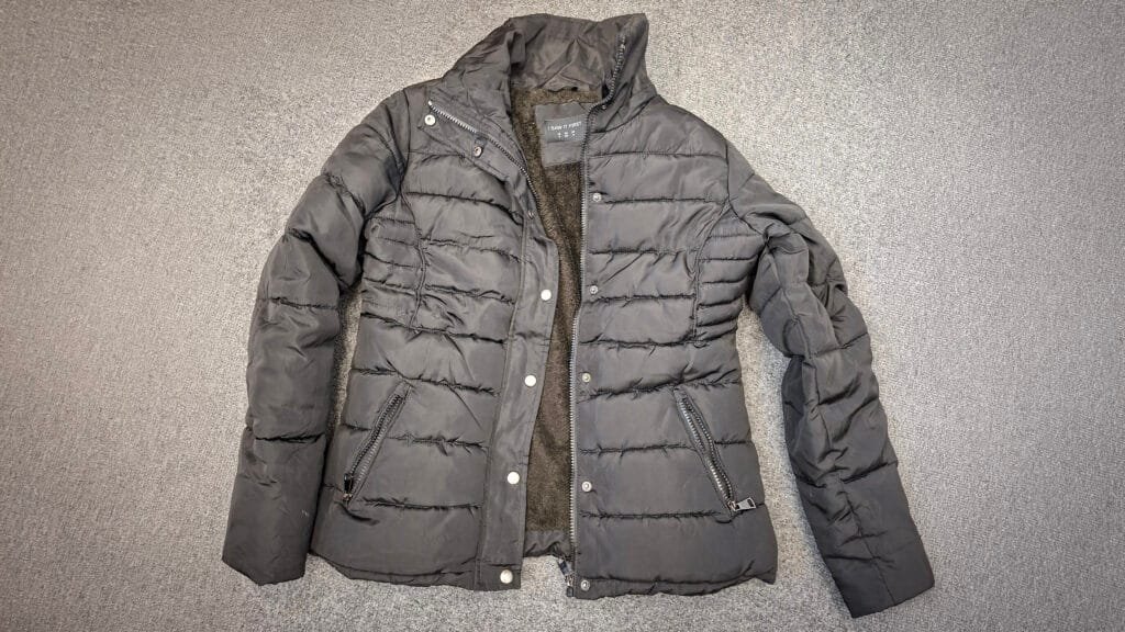 A black padded jacket with a zip and button fastening laid flat on a grey carpet