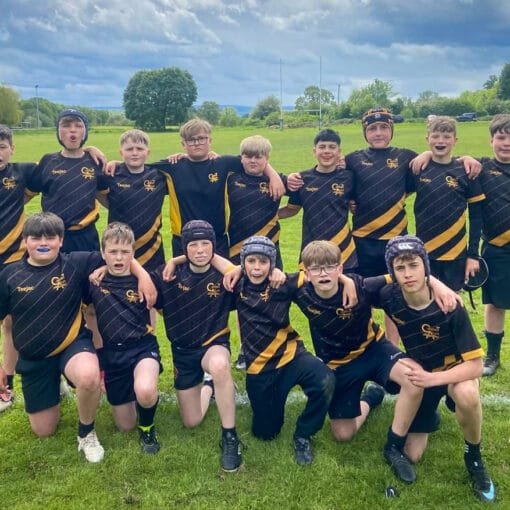 A group of students in black sports kits with yellow detailing standing and kneeling together on a grassy rugby field under a cloudy sky