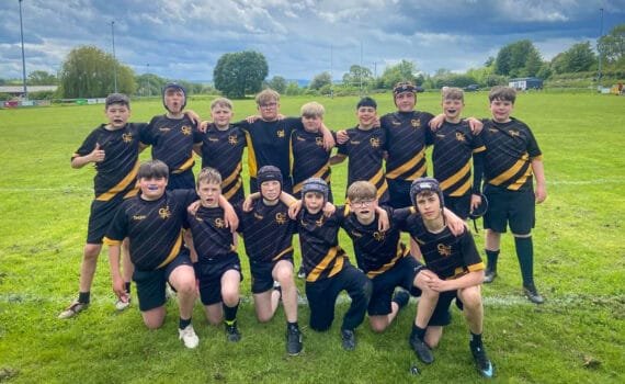 A group of students in black sports kits with yellow detailing standing and kneeling together on a grassy rugby field under a cloudy sky