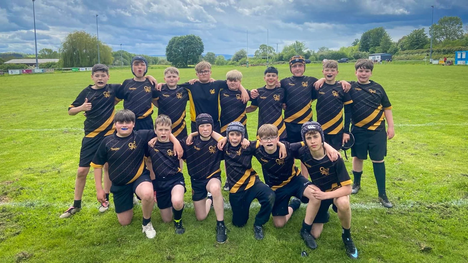 A group of students in black sports kits with yellow detailing standing and kneeling together on a grassy rugby field under a cloudy sky