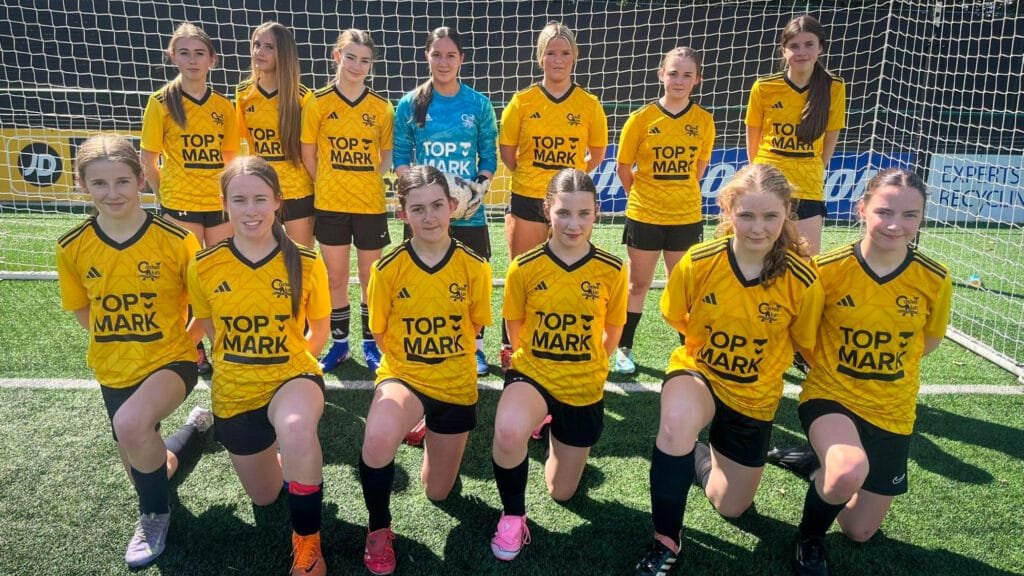 A group of students in yellow football kits posing in two rows in front of a goal on an outdoor pitch in bright sunlight