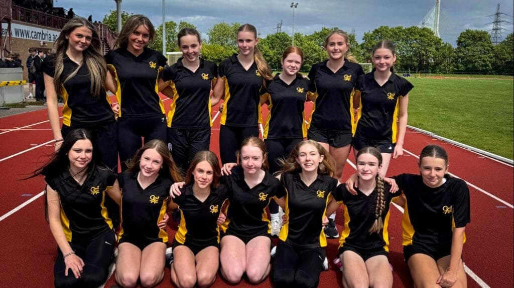 A group of students in black sports kits posing together on a running track, with stadium seating and trees in the background