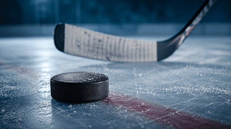 Black ice hockey puck resting on a scratched, frosty rink surface, with a taped hockey stick blade blurred in the background and cool blue arena lighting