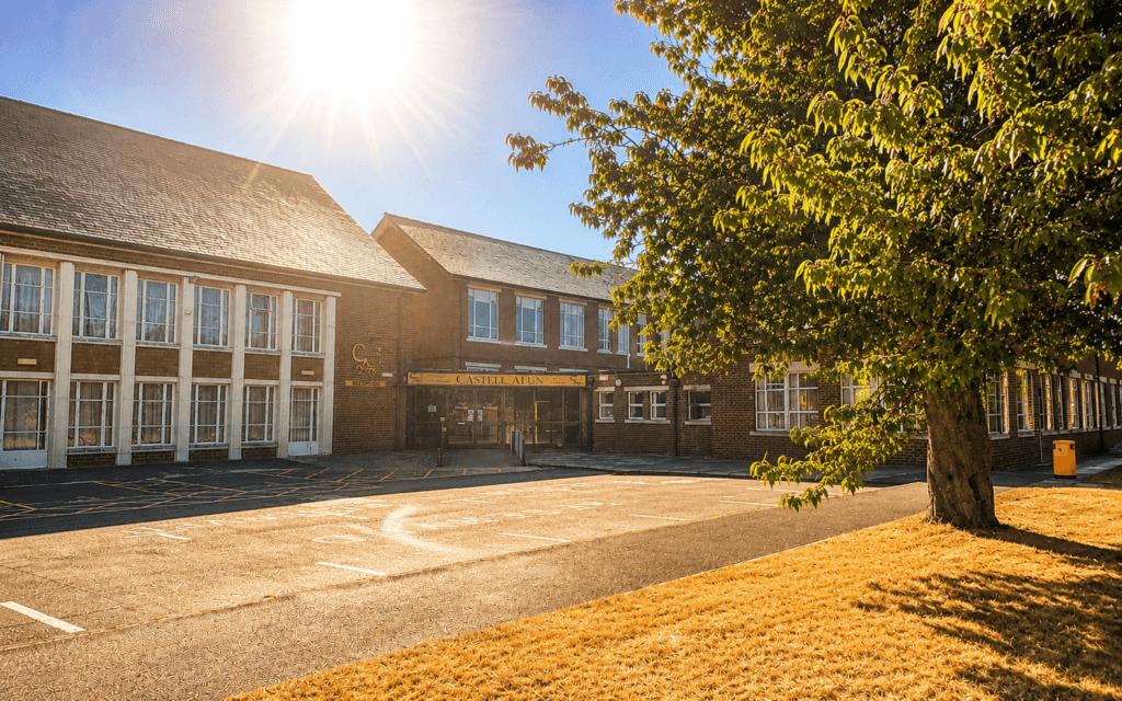 An image of the Castell Alun school building and entrance on a bright sunny day, with strong sunlight in the sky and a large tree casting shade across the front of the school.