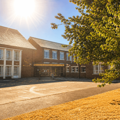 An image of the Castell Alun school building and entrance on a bright sunny day, with strong sunlight in the sky and a large tree casting shade across the front of the school.