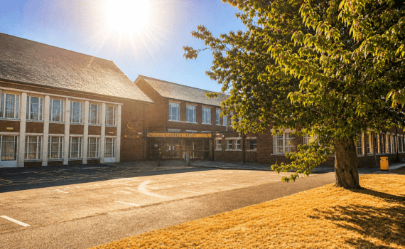 An image of the Castell Alun school building and entrance on a bright sunny day, with strong sunlight in the sky and a large tree casting shade across the front of the school.