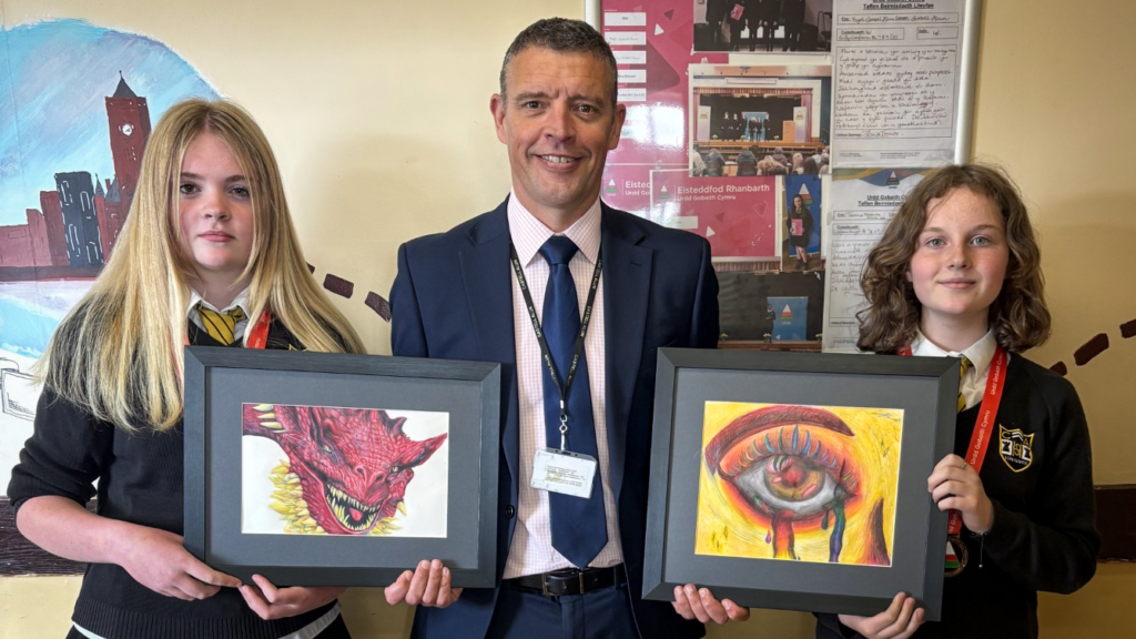 Two students in school uniform stand beside an adult in a suit, each holding framed artwork—one showing a red dragon and the other a detailed eye—against a classroom wall with posters and displays.