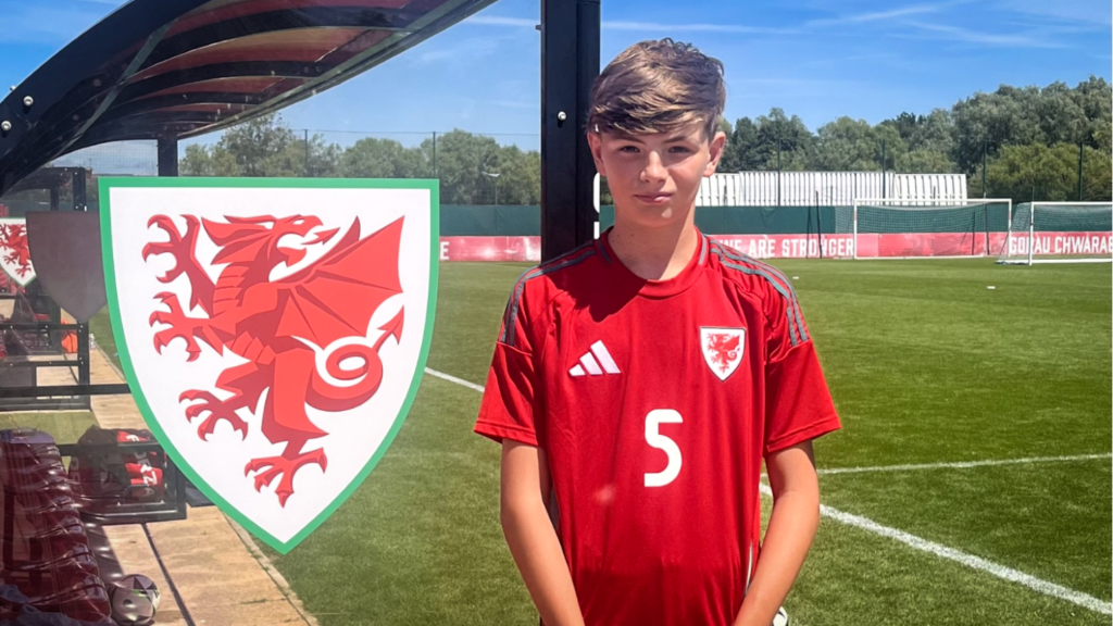 A student wearing a red Wales football kit with the number 5 stands beside a large Welsh dragon crest on a clear panel, next to a football pitch and team bench area under a sunny sky.
