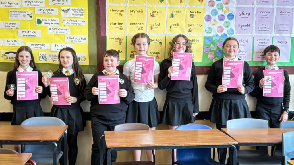 A group of students in school uniform standing in a classroom, each holding a pink certificate, with colourful Welsh language display posters on the wall behind them and desks in the foreground.