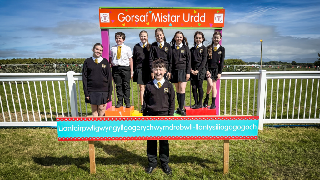A group of students in school uniform pose on a colourful display stand outdoors beneath a sign reading ‘Gorsaf Mistar Urdd,’ with a long place name sign reading ‘Llanfairpwllgwyngyllgogerychwyrndrobwllllantysiliogogogoch’ in front and a grassy field and cloudy sky behind.