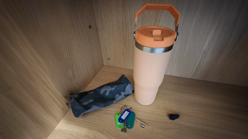 A peach-coloured insulated water bottle with a handle sits on a wooden shelf beside a grey camouflage-patterned pouch, a set of keys with coloured tags, and a small black tassel.