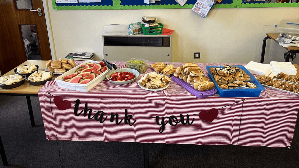 A long table covered with a red and white striped cloth displaying a variety of food including sandwiches, pastries, cakes, watermelon slices, strawberries, grapes, and plates, with a ‘thank you’ banner hanging across the front.
