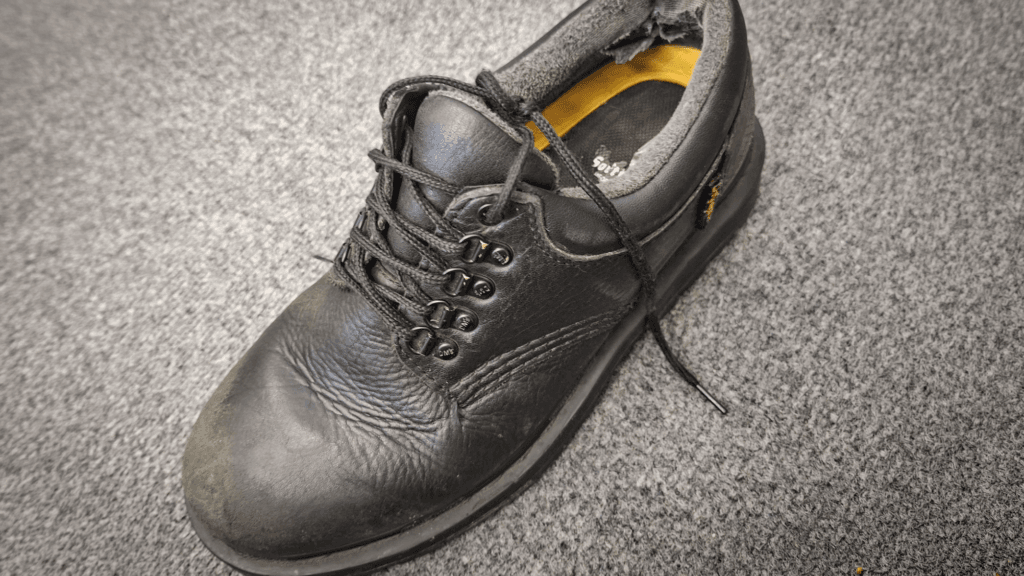 A worn black leather school shoe with laces, showing scuffs and damage around the heel and edges, resting on a grey carpet.