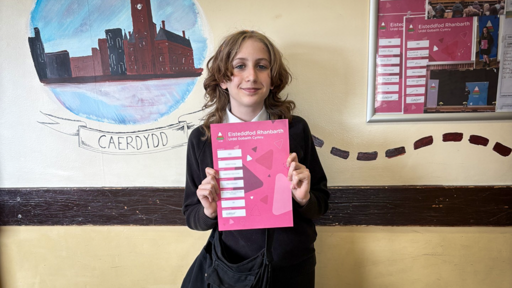 A student in school uniform stands indoors holding a pink certificate, with a painted mural of Cardiff and classroom displays behind.