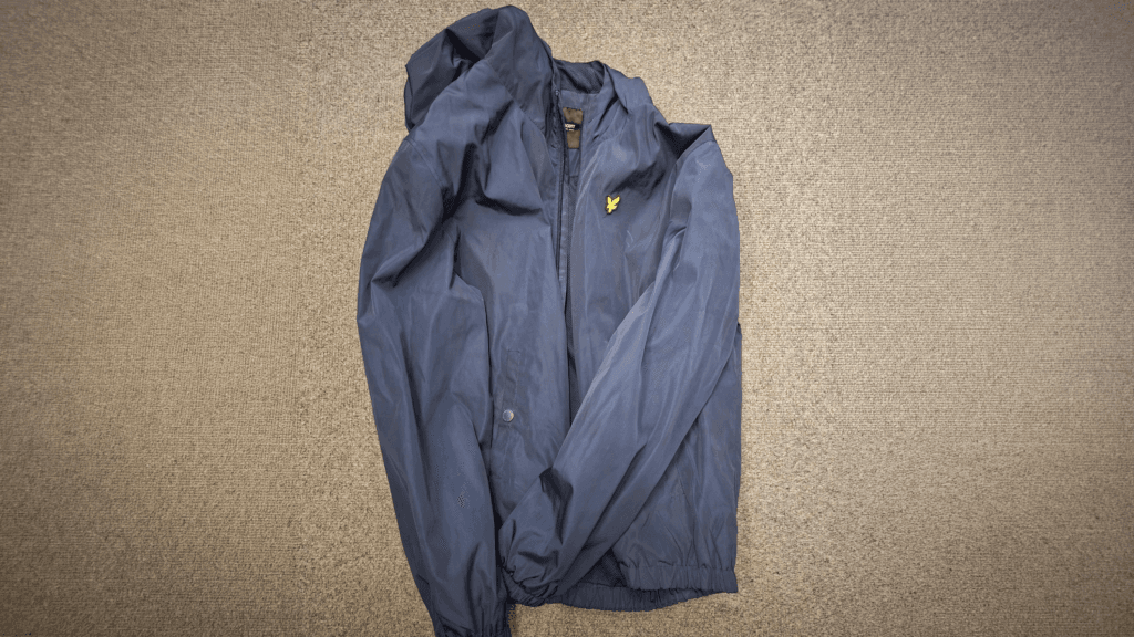 A blue hooded jacket with a zip front and small logo on the chest, laid flat on a grey carpet.