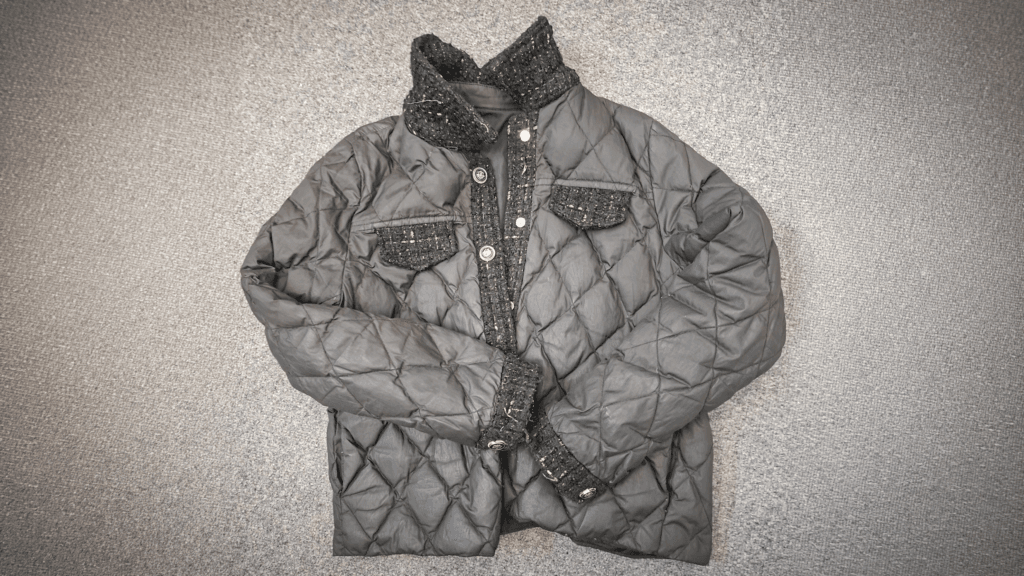 A dark grey quilted jacket with a button fastening and textured fabric panels on the collar and pockets, laid flat on a grey carpet.
