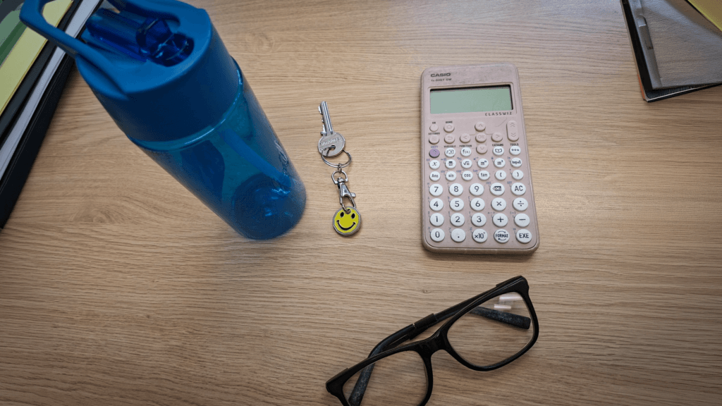 A blue water bottle, a keyring with a yellow smiley-face charm, a scientific calculator and a pair of black-framed glasses arranged on a wooden surface as part of the school’s lost property collection